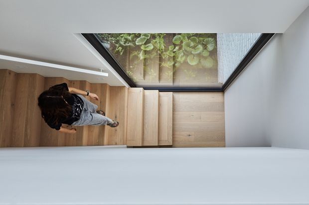 A top-down architectural view of a compact, minimalist stairway with pale timber treads and matching timber flooring. White walls and ceilings frame the stairs, while a large glazed skylight or roof opening at the landing brings in natural light and reveals greenery outside. A person is visible on the stairs.