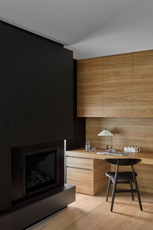 A refined compact interior featuring a built-in black fireplace wall on the left and a warm timber study nook on the right. Full-height wood veneer cabinetry, integrated drawers, a timber worktop, and a black sculptural chair create a cohesive minimalist workspace. A small pleated table lamp and books add understated detail. The room has a pale ceiling and timber flooring with a warm, restrained palette.