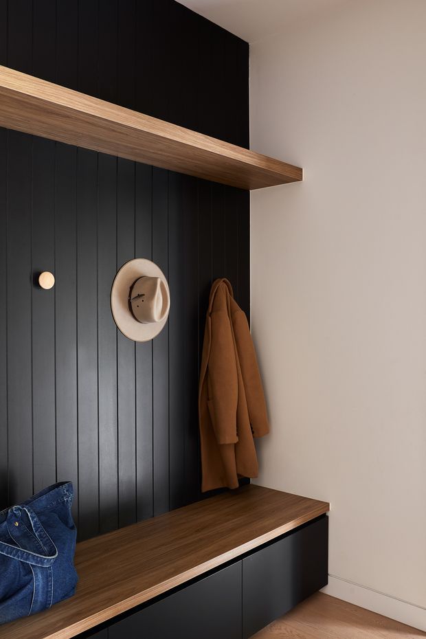 A compact, thoughtfully designed entryway with a dramatic black vertical-panel feature wall, integrated warm timber shelf and bench, and black built-in storage cabinetry. A coat and hat hang on the wall, while a denim bag rests on the bench. The space has a restrained contemporary look with warm wood accents and soft neutral surrounding walls.