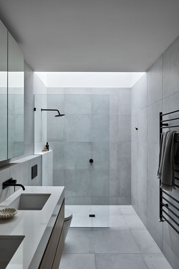 A contemporary bathroom with a walk-in tiled shower, frameless glass screen, dual integrated vanity basins, wall-mounted black tapware, large mirrored cabinet, and a black heated towel rail. Pale grey stone-look tiles and a white benchtop create a clean, minimal scheme, with a bright overhead skylight or clerestory opening illuminating the space.