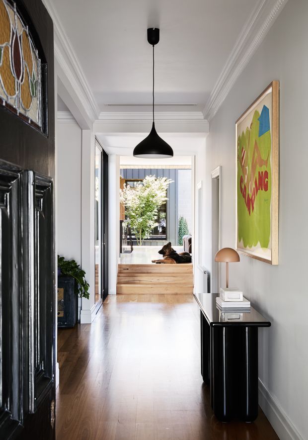 A bright, refined residential hallway with polished timber flooring, white walls and ornate decorative ceiling cornices. A black sculptural pendant light hangs centrally, while a slim black console with a brass-toned table lamp and books sits beneath colourful framed artwork. A dark decorative door with stained-glass glazing is visible on the left, and the hallway opens toward a raised living area with timber steps, glazing and a resting dog.