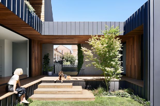 A contemporary courtyard-style residential exterior with a sheltered timber-lined outdoor living area, raised timber deck, broad steps, built-in bench seating, dark vertical cladding, and a central landscaped garden with a small tree. The open roofless courtyard brings in abundant daylight, while large glazed openings connect the interior to the outdoor space.