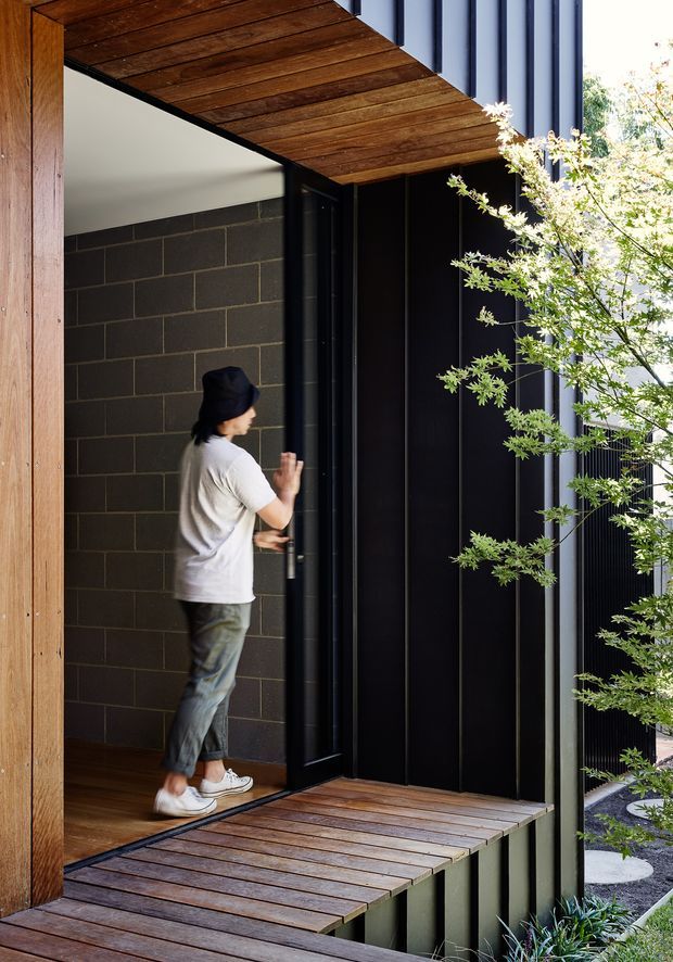 A contemporary residential entrance with a deep timber-lined overhead canopy and matching timber deck threshold. A person is opening a tall black door set within a dark vertical-clad facade, beside a charcoal masonry wall. Slender planting and a paved garden edge frame the entry, creating a strong contrast between warm wood, black cladding, and greenery.