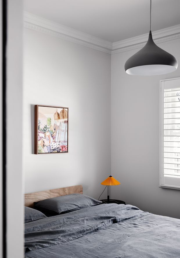 A refined bedroom viewed from the doorway, featuring a low timber headboard, charcoal-grey rumpled bedding, pale grey walls, ornate white cornice moulding, a framed artwork, a large black sculptural pendant light, and a small orange pleated bedside lamp. White plantation shutters provide filtered daylight at the window.