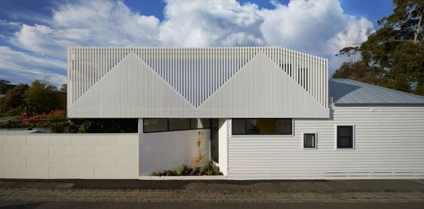Exterior view of a contemporary low-rise house with a distinctive white vertical slatted upper facade forming repeated triangular peaks. The lower level combines white horizontal weatherboard cladding, pale masonry walling, dark-framed windows, and a grey metal pitched roof. A narrow planted entry court sits beneath the projecting upper volume, with trees and a partly cloudy sky in the background.