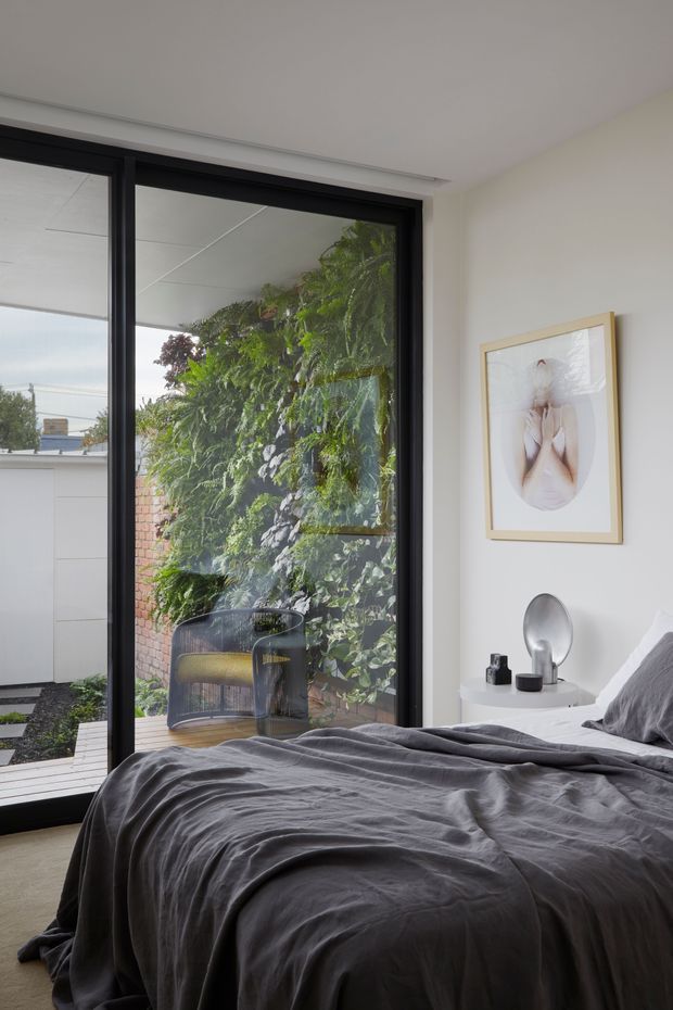 A calm, contemporary bedroom with a low-profile bed dressed in dark grey textured bedding. The room has white walls and ceiling, slim black-framed floor-to-ceiling glazing opening toward a leafy courtyard, and a small white round bedside table with minimal decorative objects. A light timber-framed artwork hangs above the table. The glazing and courtyard planting bring abundant greenery and natural light into the room.
