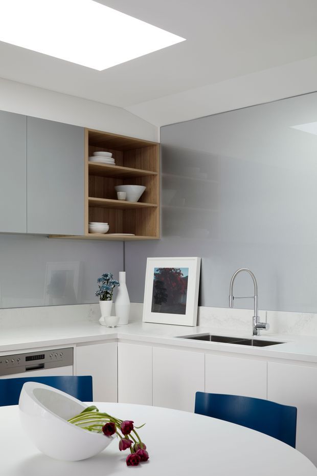 A bright, compact contemporary kitchen and dining area with white flat-panel cabinetry, a grey glass splashback, white benchtops, a stainless steel sink and gooseneck tap, and an open timber display shelf. A large ceiling skylight provides strong natural illumination. The foreground contains a white round dining table with blue dining chairs and simple decorative items.