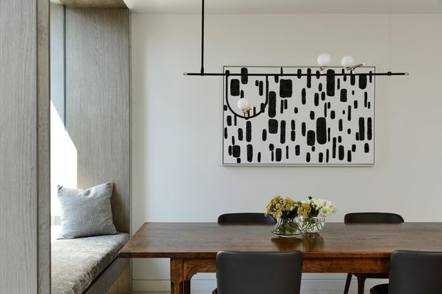 A refined dining area with a long dark timber dining table and dark upholstered chairs, positioned beside a built-in window seat with a pale cushion. Light textured timber wall panelling frames the window, while a minimalist linear pendant light hangs above the table. A monochrome abstract artwork and a small floral arrangement add restrained decorative detail. The space is naturally lit with a neutral, warm palette.