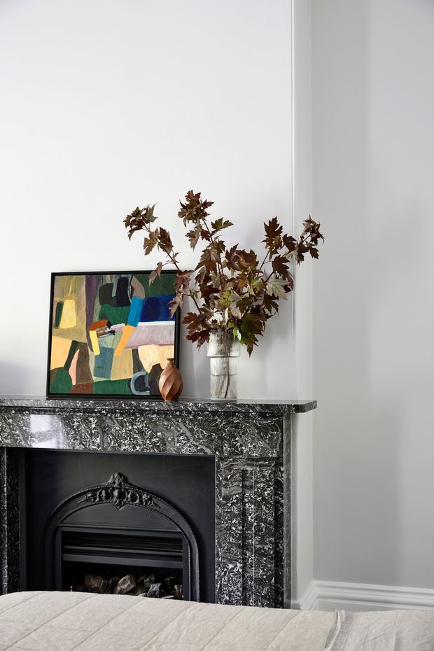 A close-up interior view of a bedroom with a traditional black fireplace and ornate dark marble surround. A colourful abstract artwork, glass vase with leafy branches, and small decorative vessel sit on the mantel. White walls with decorative skirting and a neutral bed in the foreground create a refined, classic setting.