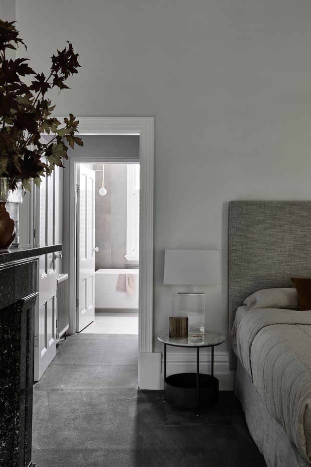 A refined bedroom viewed toward an open doorway leading to a bathroom. The room features a grey upholstered bedhead, layered neutral bedding, a round black metal and glass bedside table, and a dark stone fireplace or console with a tall leafy arrangement. Grey carpet, pale grey walls, white traditional door and window trims, and soft neutral furnishings create a calm, monochromatic interior.