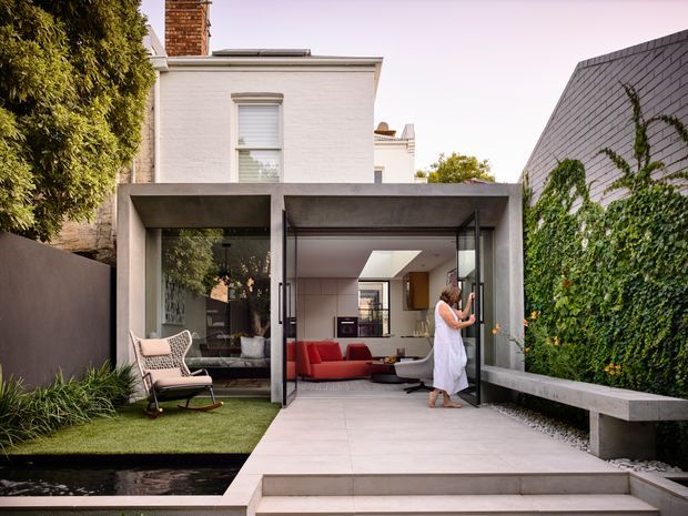 A contemporary rear extension to a traditional painted brick residence, viewed from a landscaped courtyard. The extension has a strong concrete frame, extensive black-framed glazing, a large open glass door, and a flat roof with a skylight. Inside is a compact living area with red seating and pale built-in cabinetry. The courtyard includes pale tiled paving, raised steps, a narrow reflecting pool, artificial or manicured lawn, a built-in concrete bench, and a vine-covered masonry wall.