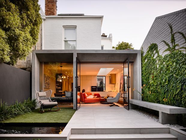 A contemporary rear extension to a narrow urban residence, viewed from an outdoor courtyard. The original white-painted masonry upper storey sits above a strongly framed glazed ground-floor addition with large opening glass doors. Inside is a warm-toned open-plan living area with a vivid red sofa, while the courtyard features pale paving, a raised grass platform, a narrow reflecting pool, a dark boundary wall, a concrete bench, and an ivy-covered masonry wall.