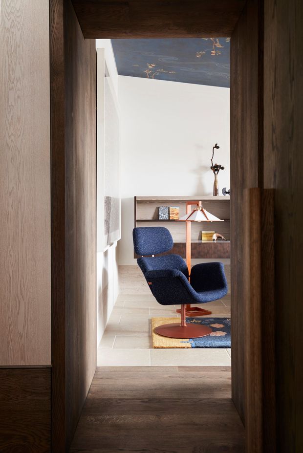 A refined interior view through a dark timber-framed doorway into a compact study or reading nook. The room features a sculptural navy lounge chair, mid-century floor lamp, built-in timber shelving, a patterned rug, pale tiled flooring, white walls, and a decorative blue ceiling with delicate botanical detailing. The composition emphasises layered timber joinery and a carefully curated, warm modernist interior.