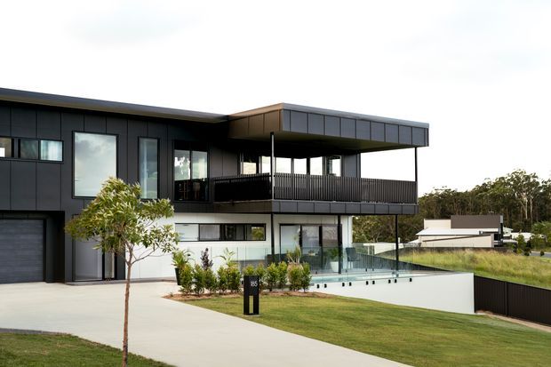 Contemporary multi-level residence with dark cladding, broad horizontal rooflines, extensive glazing, a projecting upper-level balcony, and a glass-edged swimming pool. The landscaped frontage includes a concrete driveway, lawn, young trees, and a dark perimeter fence.