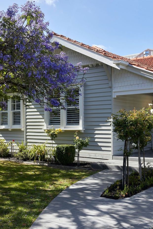 Exterior view of a light-coloured, single-storey residential house with horizontal weatherboard cladding, white trim, louvred windows, a covered entry or carport opening, and a terracotta tiled pitched roof. A paved walkway leads through a landscaped front garden with mature flowering trees and clipped planting.