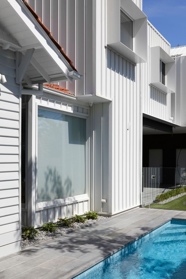 Bright exterior view of a contemporary residential building beside a swimming pool. The facade combines white vertical ribbed cladding with sections of horizontal weatherboard siding, projecting window surrounds, large glazed openings, and a covered garage or outdoor area. A pale tiled pool deck and landscaped planting line the pool edge, with strong sunlight and crisp shadows creating a clean, modern appearance.