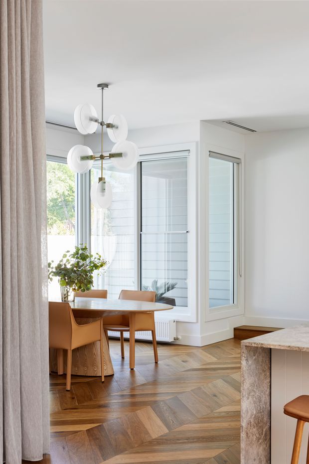 Bright, refined open-plan dining area adjoining a kitchen, with a round timber dining table and tan leather chairs beside large corner windows. The room features herringbone timber flooring, white walls and built-in window surrounds, a sculptural globe pendant light, full-height curtains, and a stone kitchen island partially visible in the foreground.