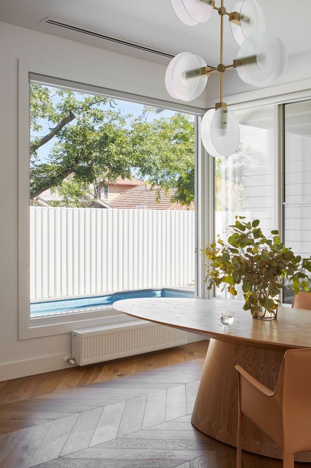 Bright, refined dining area with a sculptural multi-arm pendant light, warm timber dining table and chairs, white walls, and a large picture window overlooking a compact swimming pool and neighbouring greenery. The room features herringbone timber flooring, a white radiator beneath the window, and glazed doors or windows along the right side.