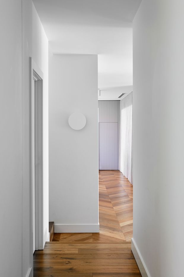 A bright, minimalist residential hallway viewed toward a deeper corridor. The space has crisp white walls and ceiling, a simple circular wall light, white vertical detailing or curtains along the right side, and warm timber flooring laid in a geometric chevron or herringbone pattern. A doorway and steps are visible on the left, with clean, understated architectural lines throughout.