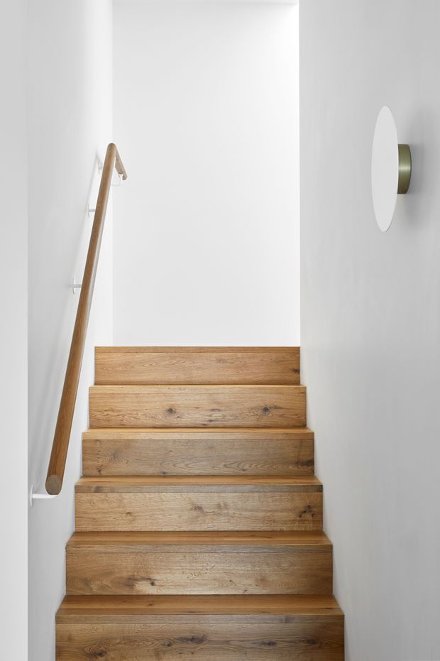 A narrow, minimalist interior stairway with solid warm-toned timber treads and risers, white painted walls, a simple timber handrail on the left, and a rounded wall sconce with a metallic backing on the right. The stair rises toward a bright white landing.