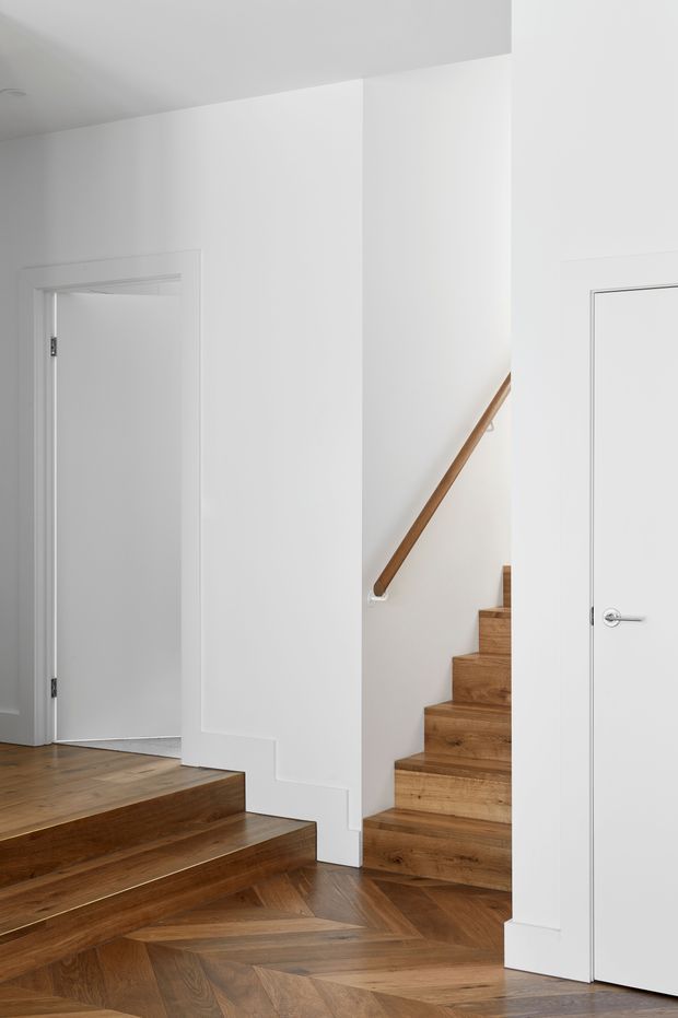 A bright, minimalist interior stairway and landing with white walls, concealed white doors, warm timber stairs, and a geometric herringbone timber floor. Two stair flights meet around a central white wall, with a simple timber handrail ascending along the right wall.