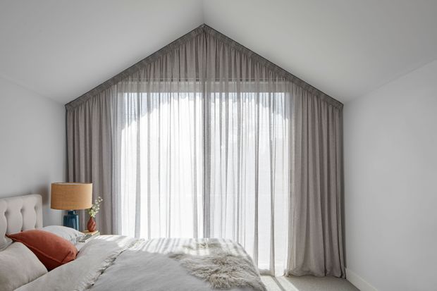 A bright, softly styled bedroom with a large peaked window dressed in full-height sheer and grey drapery. The bed occupies the foreground with layered neutral bedding and a muted rust accent cushion, while a bedside table and warm table lamp sit to the left. The room has plain white walls, a simple sloping ceiling, and a calm monochromatic palette.