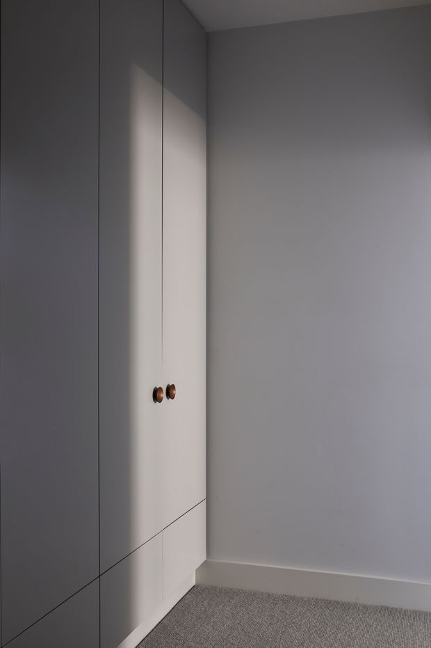 A narrow, softly lit built-in storage area with tall flush-panel cabinetry along the left wall, featuring small dark metallic or timber knobs. The surrounding walls and ceiling are painted pale grey, with a light grey carpeted floor and simple white skirting. The restrained palette and clean lines create a quiet, minimalist interior.