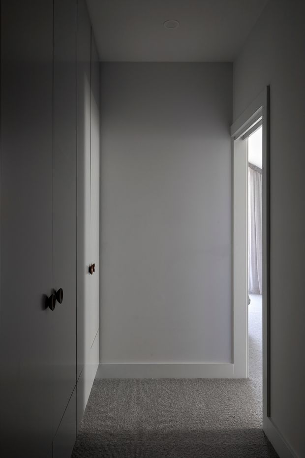 A narrow, dimly lit residential passage or wardrobe area with full-height grey built-in cabinetry on the left, dark round pulls, pale grey walls, white trim, and a grey carpeted floor. An open doorway on the right leads into a brighter adjoining room with a curtained window. The restrained palette and flush joinery create a quiet, minimalist atmosphere.