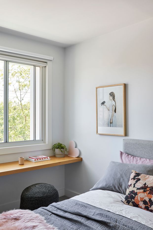 A bright, compact bedroom with pale grey walls, a large window, and a wall-mounted timber desk or shelf beneath the glazing. The room includes a grey upholstered bedhead, layered grey and white bedding, blush and patterned cushions, a black knitted ottoman, a small potted plant, and framed artwork. The overall look is calm, contemporary, and softly styled with natural light.