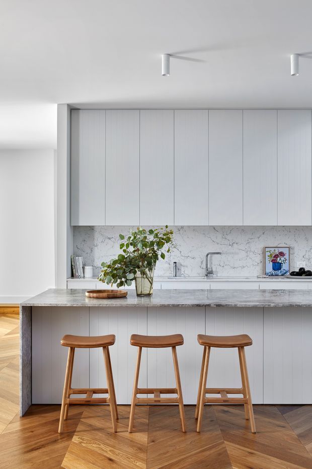 A bright, refined contemporary kitchen with a large island and three timber bar stools. The kitchen features floor-to-ceiling pale vertical-panel cabinetry, a white marble-look veined splashback and benchtop, integrated appliances, and a sleek stainless-steel tap. Warm herringbone timber flooring contrasts with the predominantly white and light-grey palette. Minimal white track lighting and sparse styling create a clean, calm atmosphere.