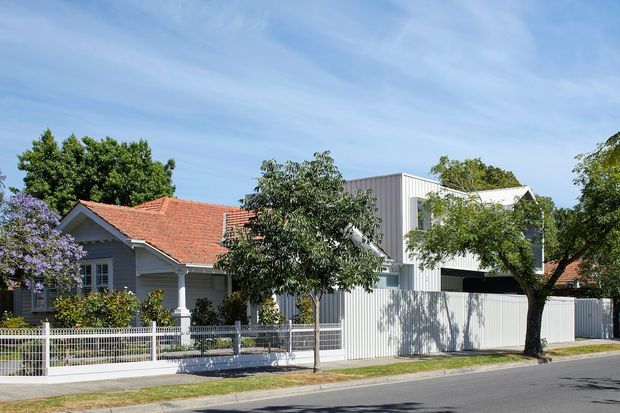 Street-facing view of a residential property combining an older single-storey weatherboard house with a red tiled pitched roof and a newer two-storey extension clad in pale vertical panels. The home sits behind a low metal fence with mature garden trees and a paved footpath, in a suburban setting under a bright blue sky.