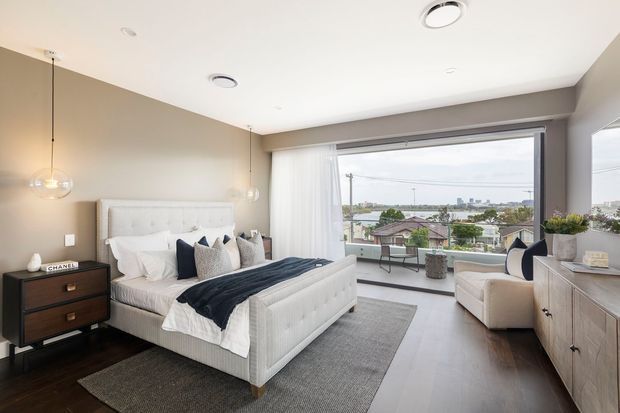Bright, contemporary bedroom with a large upholstered bed, layered neutral bedding, dark navy accents, bedside furniture, pendant lights, and a wide glazed opening to a balcony with an urban outlook. A low timber sideboard and seating area complete the refined, airy room.