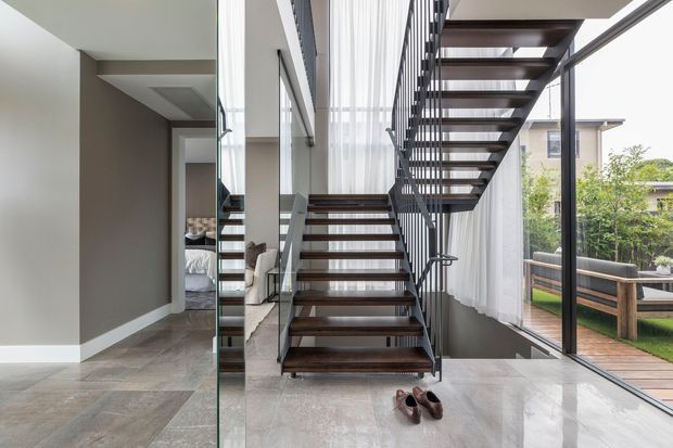 A bright contemporary interior centered on an open-tread floating staircase with dark timber steps, slim dark metal stringers and railings, and a glass balustrade. Floor-to-ceiling glazing with sheer curtains opens toward a timber deck and landscaped garden. A bedroom is visible through the opening on the left, while polished large-format stone-look flooring and neutral walls create a clean, airy feel.