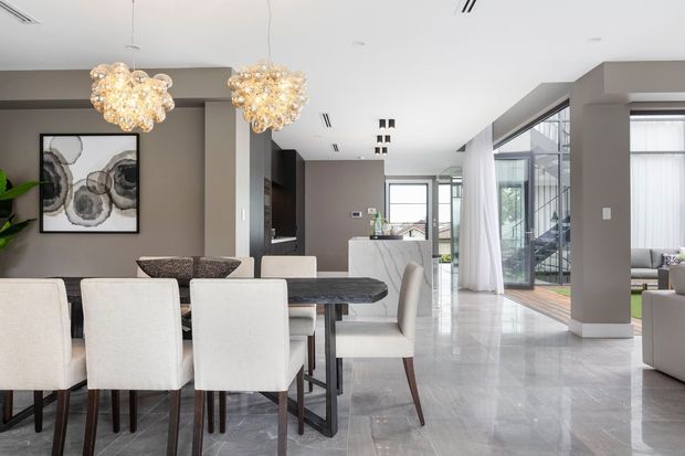 A sophisticated open-plan dining and living area with a contemporary kitchen beyond. The space features a large dark dining table with upholstered cream dining chairs, taupe-grey walls, polished light-grey stone flooring, sculptural clustered pendant lights, and expansive floor-to-ceiling glazing with sheer curtains. A staircase and additional living seating are visible toward the rear, creating a bright, refined interior.