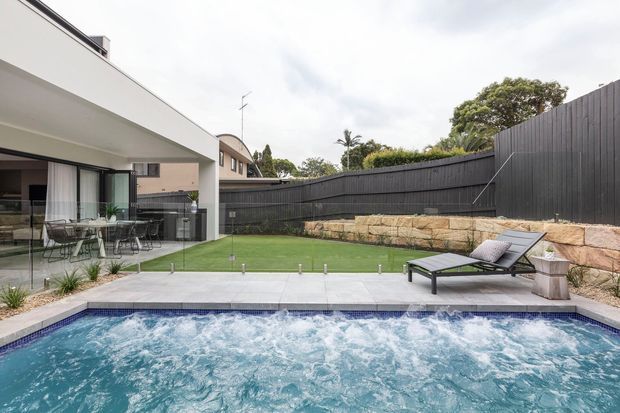 Contemporary backyard pool area with a bubbling rectangular swimming pool in the foreground, pale tiled coping and paving, and two sun loungers. A covered outdoor living and dining area with frameless glazing sits to the left, opening onto a small artificial-turf courtyard. Dark vertical fencing, a sandstone retaining wall, and established planting enclose the space.