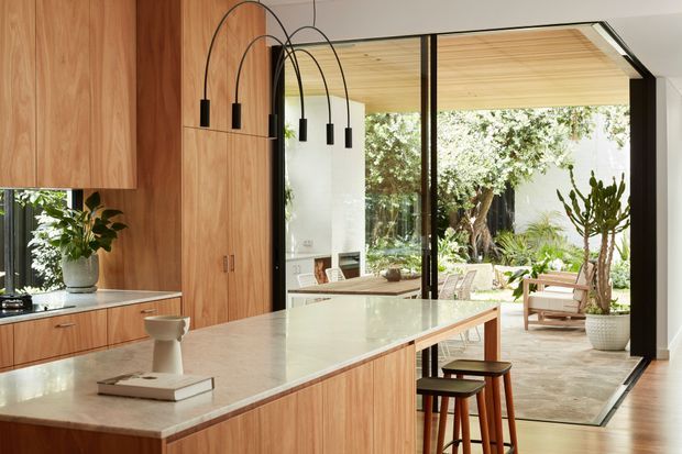 A bright contemporary kitchen with extensive warm timber cabinetry and a large pale stone island. Black arched pendant lights hang above the island, which has seating for several people. Full-height black-framed glazed doors open directly to a furnished outdoor terrace and lush garden, creating a strong indoor–outdoor connection.