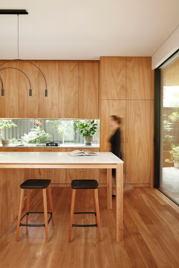 Warm contemporary kitchen interior with extensive light timber cabinetry, a white island benchtop with two black stools, timber flooring, integrated cooktop and sink, arched pendant lights, and large glazed openings to greenery outside.