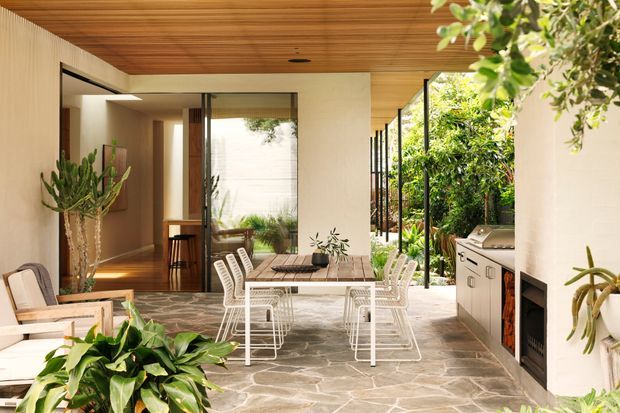 A refined, open-air courtyard dining and outdoor kitchen area connected to a contemporary home. The space features a timber-lined ceiling, pale rendered walls, natural irregular stone paving, extensive glazing and slim dark frames, a built-in barbecue and cabinetry, a dining table with white wire-frame chairs, and abundant greenery surrounding the partially enclosed terrace.