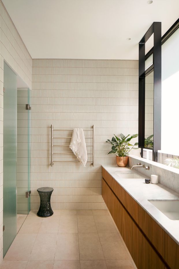 Bright contemporary bathroom with a long timber vanity, pale stone benchtop and integrated basins, large black-framed mirror, full-height narrow cream wall tiles, and matching pale floor tiles. A frameless glass shower enclosure is visible on the left, with a brass heated towel rail and indoor plant adding warmth. The room is naturally lit through large glazing on the right.