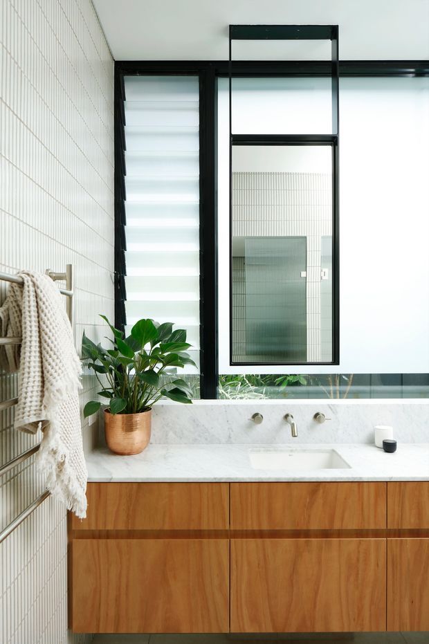 A bright contemporary bathroom featuring a timber floating vanity with integrated white marble-look benchtop and basin, brushed metal wall-mounted tapware, black-framed louvre and awning windows, and pale vertically ribbed wall tiles. A towel rail with a knitted towel and a potted plant add warmth and texture.