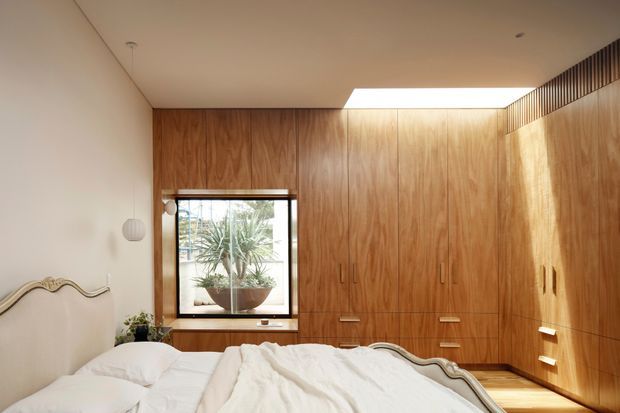 A warm, compact bedroom with a built-in timber wardrobe spanning the rear and right walls. The room includes a bed with a curved upholstered headboard, white bedding, a small window with a potted plant, recessed lighting, and a high-level skylight or clerestory opening that brings in natural light. The joinery has vertical grain timber veneer and integrated handles, creating a calm, refined built-in interior.