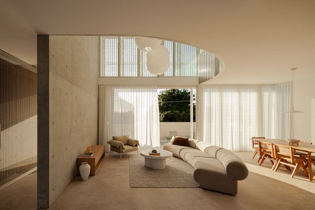 A warm, light-filled open-plan living and dining space with a sculptural curved sectional sofa, rounded coffee table, timber sideboard, and wood dining table with chairs. The room features pale polished concrete walls, a large curved clerestory opening, full-height sheer curtains, vertical timber screening, and distinctive suspended pendant lights. Floor-to-ceiling glazing opens toward a leafy exterior, while the restrained palette and rounded furniture create a refined contemporary atmosphere.