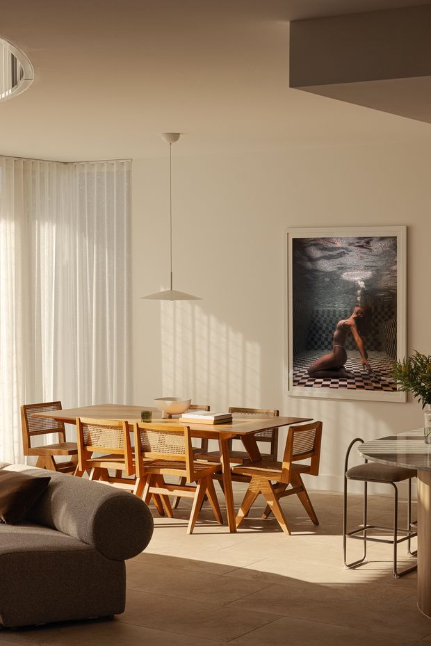 Warm, sunlit open-plan dining and living area with a mid-century-inspired timber dining table and cane-back chairs. A neutral upholstered sofa occupies the foreground, while a pendant light, sheer vertical curtains, framed artwork, and a small side table with bar stool complete the room. The space has pale walls, large light stone or tile flooring, and strong afternoon shadows.