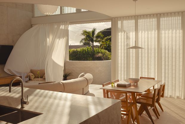 Warm, sunlit open-plan interior combining a kitchen, dining area and living space. A pale stone kitchen island with undermount sink occupies the foreground, while a timber dining table and chairs sit beside full-height sheer curtains and glazed openings. The living area includes a curved cream sofa, soft white drapery, pale masonry surfaces and a small courtyard visible beyond. The space has a refined contemporary character with strong indoor-outdoor connection and filtered natural light.