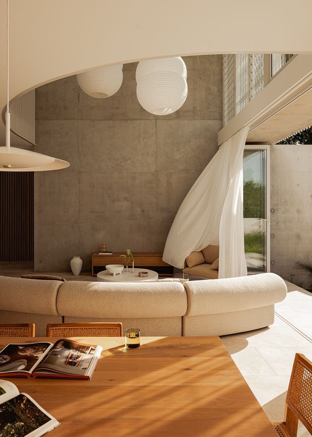 Warm, sunlit contemporary living and dining interior with a sculptural curved cream sofa in the foreground, a timber dining table and woven chairs, a low timber media console, and a softly curtained seating nook. The room features a raw exposed concrete wall, pale tiled flooring, expansive glazing, curved ceiling detailing, and large white paper pendant lights. The palette is earthy, neutral, and tactile with strong indoor-outdoor light.