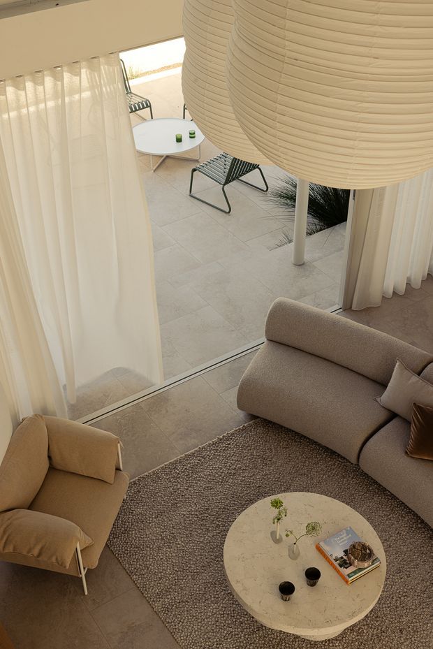 A warm, softly styled living room viewed from above, opening directly onto a light-toned tiled patio. The room features a curved beige sofa, matching upholstered armchair, textured neutral rug, round pale stone coffee table, and sheer floor-to-ceiling curtains. Outside, a round table, outdoor lounge chairs, sculptural oversized pendant-like shade elements, and sparse ornamental planting create a minimalist courtyard setting.