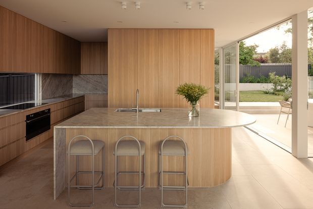 A refined open-plan kitchen with a large curved-end island, integrated sink, three stools, extensive vertical timber cabinetry, and a stone-look benchtop. Dark appliances and a veined stone splashback contrast with the warm timber joinery. Full-height glazed doors open to a bright garden courtyard and outdoor seating.