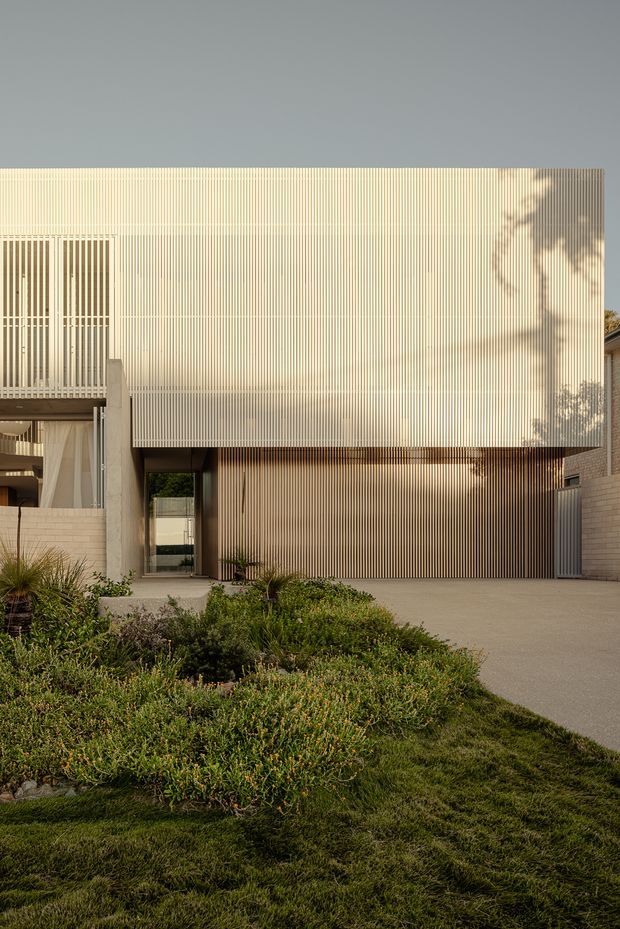 A modern residential facade viewed from a landscaped front garden. The building features a large projecting upper volume clad in closely spaced vertical light-coloured slats, with a darker timber-toned slatted wall beneath, recessed glazing, and a concrete-framed balcony or entry element. Low native-style planting and lawn occupy the foreground, while a paved driveway or forecourt leads to the building.