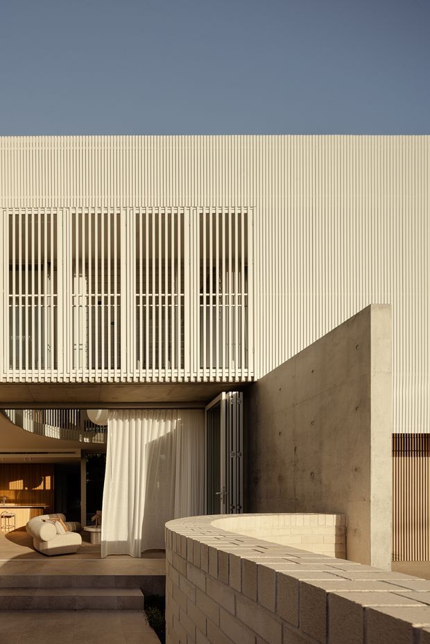 A contemporary residential courtyard facade viewed from outside, with a pale vertically ribbed upper facade, extensive white vertical screening over glazed openings, exposed concrete walls, and a curved pale brick boundary wall in the foreground. An open living area with curtains, seating, and warm timber finishes is visible through the lower-level opening. Warm late-day sunlight creates strong shadows and a refined, textural composition.