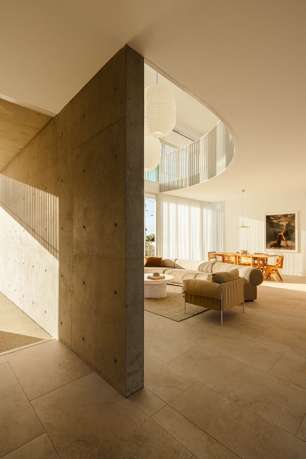 A warm, sunlit contemporary living and dining space with a sculptural oval ceiling opening overlooking an upper-level void. A monolithic exposed concrete wall divides the circulation area from the furnished room. The interior features pale large-format tiled flooring, full-height sheer window coverings, curved cream sofas and armchairs, a low round coffee table, a timber dining table with matching chairs, and suspended ribbed pendant lights.